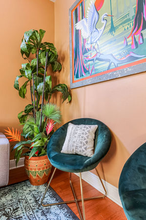 Cozy living room corner with a teal velvet accent chair and patterned pillow, terracotta pot of tropical plants, colorful heron wall art on a warm peach wall, hardwood floor and patterned rug.