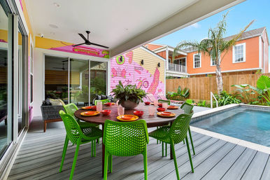 Vibrant covered backyard pool patio with a round wooden dining table set with colorful plates and bowls, lime-green chairs, potted centerpiece, sliding glass doors, a pink mural wall, tropical plants and a rectangular swimming pool.