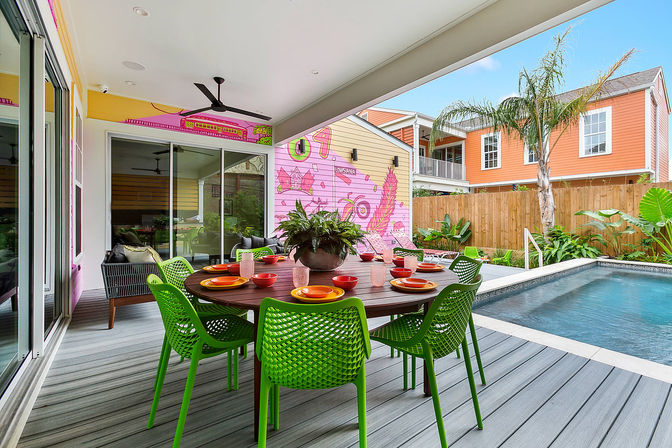 Vibrant covered backyard pool patio with a round wooden dining table set with colorful plates and bowls, lime-green chairs, potted centerpiece, sliding glass doors, a pink mural wall, tropical plants and a rectangular swimming pool.