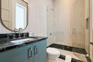 Sleek modern residential bathroom with black marble countertop, teal vanity, oval mirror and chrome faucet, white toilet and glass-enclosed walk-in shower with beige tile
