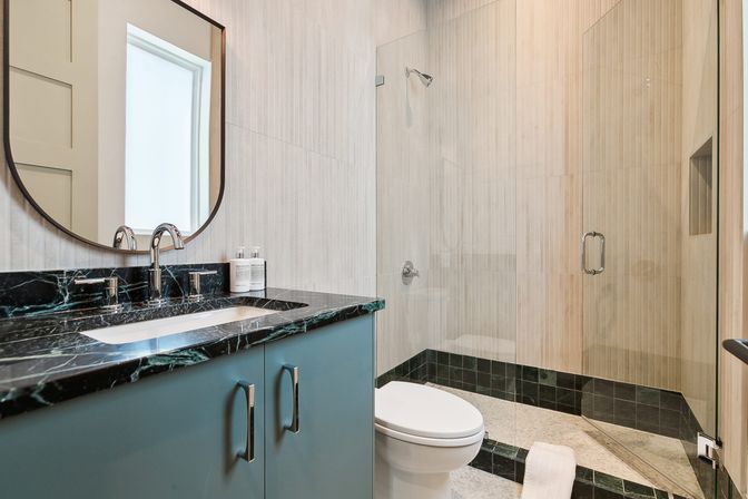 Sleek modern residential bathroom with black marble countertop, teal vanity, oval mirror and chrome faucet, white toilet and glass-enclosed walk-in shower with beige tile