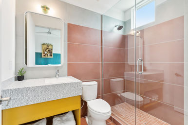 Bright sunlit modern bathroom with pink-tiled walk-in shower and glass door, terrazzo countertop on a bold yellow vanity, white toilet, small potted plant and high window.
