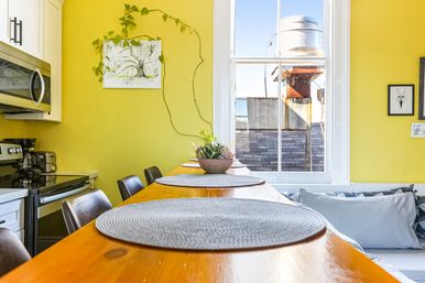 Sunny urban kitchen with bright yellow walls and a wooden breakfast bar set with round gray placemats, black stools, a succulent centerpiece, vine-accented wall art, and a window overlooking a rooftop and metal water tank.