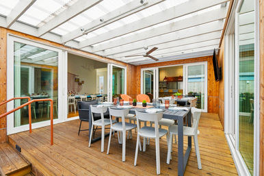 Sunlit covered patio on a wooden deck with a long outdoor dining table, white chairs, potted succulents under a translucent pergola and ceiling fan, with sliding glass doors to a modern kitchen.