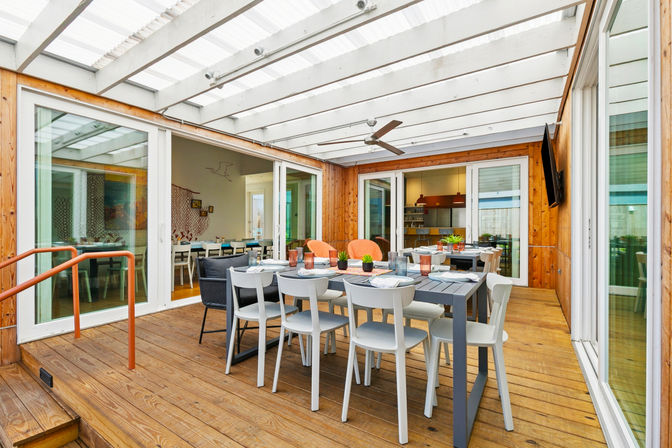 Sunlit covered patio on a wooden deck with a long outdoor dining table, white chairs, potted succulents under a translucent pergola and ceiling fan, with sliding glass doors to a modern kitchen.