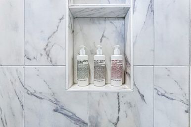 Three white pump bottles of toiletries arranged in a recessed marble shower niche set into white and gray veined tiles