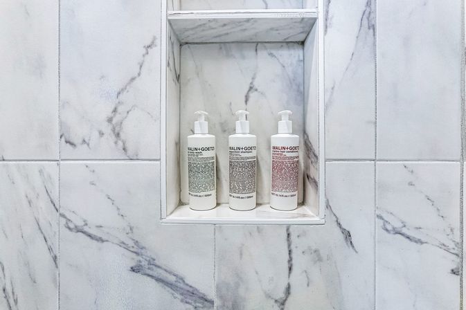 Three white pump bottles of toiletries arranged in a recessed marble shower niche set into white and gray veined tiles