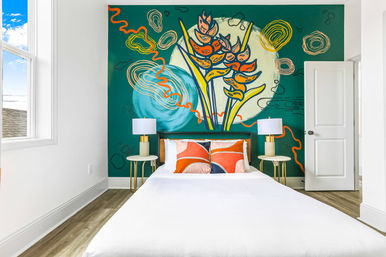 Bright modern bedroom with bold teal mural of abstract tropical flowers, white bed, orange accent pillows, gold bedside lamps and sunlit window