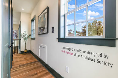 Sunlit contemporary hallway in a modern home with warm hardwood floors, green-trim window showing blue sky and trees, abstract framed artwork, potted plant, and wall lettering referencing an assemblage and installation by the Afrofuture Society.