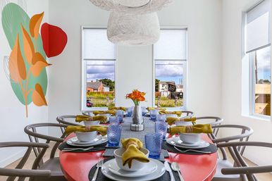 Bright modern dining room with a red oval table set for eight — white dishes, textured blue glasses and mustard-yellow napkins, curved wooden chairs, woven pendant lights, abstract leaf wall mural and a vase of orange flowers under large windows with a suburban neighborhood view.