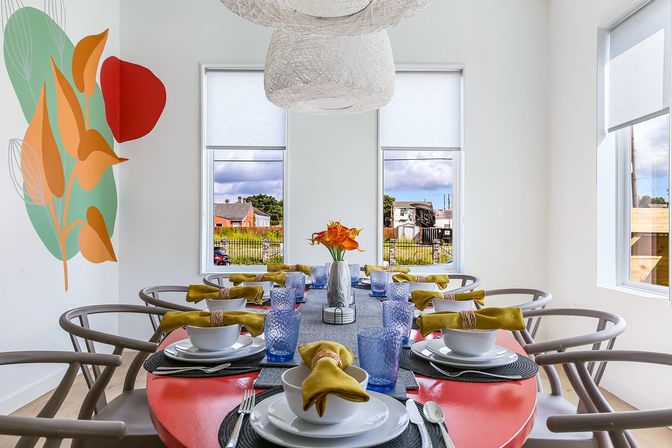 Bright modern dining room with a red oval table set for eight — white dishes, textured blue glasses and mustard-yellow napkins, curved wooden chairs, woven pendant lights, abstract leaf wall mural and a vase of orange flowers under large windows with a suburban neighborhood view.