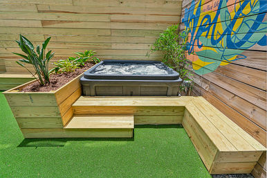 Cozy backyard patio with bubbling hot tub set into built-in wooden planter and bench, bright artificial turf and colorful mural on a wood fence.