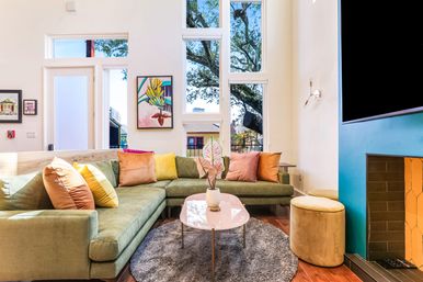 Cheerful modern living room with floor-to-ceiling windows, green L-shaped sectional sofa, colorful throw pillows, blush round coffee table on a gray rug, yellow pouf and fireplace accent.