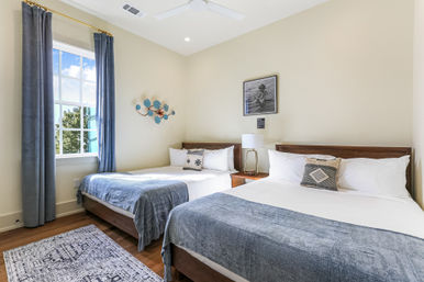 Sunny modern guest bedroom with two beds dressed in white linens and denim-blue throws, wooden headboards, bedside table with lamp, window framed by blue curtains, decorative wall art and patterned rug.
