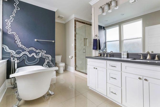 Bright modern master bathroom with white clawfoot tub, navy abstract mural accent wall, glass walk-in shower, glossy tile floor, and white double-sink vanity with chrome fixtures