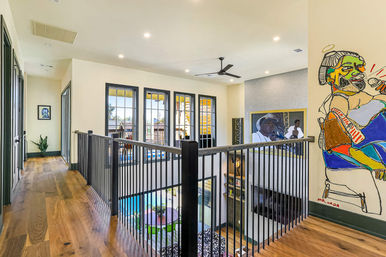 Sunlit modern upstairs hallway in an open-concept home with warm hardwood floors, black metal balcony railing overlooking a two-story living area and pool, large multi-pane windows, ceiling fan, recessed lights, and colorful wall art and framed portrait.