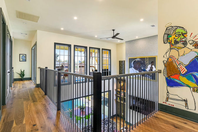 Sunlit modern upstairs hallway in an open-concept home with warm hardwood floors, black metal balcony railing overlooking a two-story living area and pool, large multi-pane windows, ceiling fan, recessed lights, and colorful wall art and framed portrait.