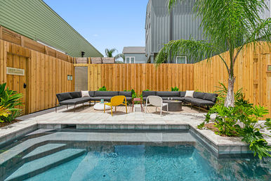 Sunny backyard with small rectangular pool, tiled patio, modern curved black sectional sofa, yellow accent chair, palm trees and wooden privacy fence.