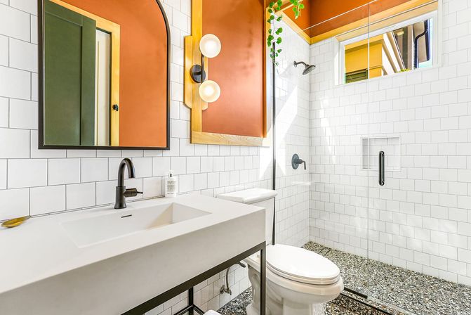 Sunlit modern bathroom with white subway tile, glass shower, terrazzo pebble floor, black fixtures, orange accent wall, arched mirror and concrete sink.