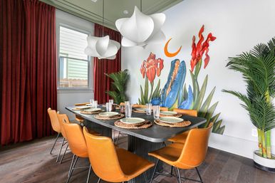 Modern dining room with a black rectangular table set for eight, orange leather chairs, woven placemats, sculptural white pendant lights, deep red curtains, tropical potted plants and a vibrant wall mural of oversized flowers and a crescent moon.
