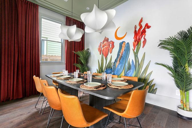 Modern dining room with a black rectangular table set for eight, orange leather chairs, woven placemats, sculptural white pendant lights, deep red curtains, tropical potted plants and a vibrant wall mural of oversized flowers and a crescent moon.