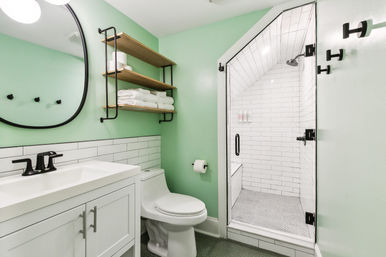 Modern mint-green bathroom with white vanity and round mirror, black fixtures, open wood shelves stacked with towels, and a glass-door walk-in shower tiled in white subway tile.