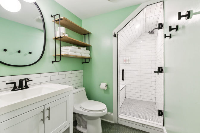 Modern mint-green bathroom with white vanity and round mirror, black fixtures, open wood shelves stacked with towels, and a glass-door walk-in shower tiled in white subway tile.