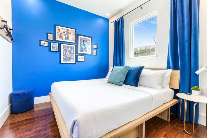 Bright modern bedroom with a bold cobalt accent wall and framed gallery art, white linens on a wooden platform bed, blue velvet curtains, teal throw pillows, sunlit window, round bedside table and hardwood floor.