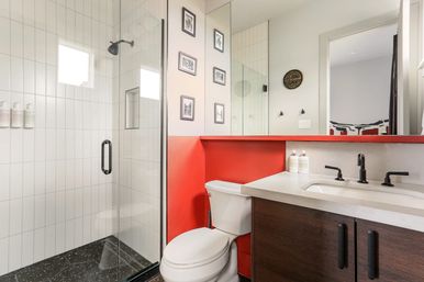 Contemporary bathroom interior with glass-enclosed white-tile shower, black fixtures, dark wood vanity with stone countertop, red accent half-wall, toilet, and large mirror.