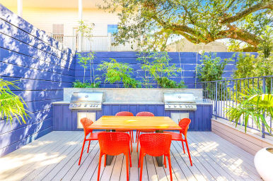 Sunny backyard deck with a wooden dining table and bright red chairs, built-in stainless-steel grills set into a blue privacy wall, bamboo planters and an overhanging oak tree — modern outdoor dining patio.