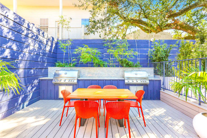 Sunny backyard deck with a wooden dining table and bright red chairs, built-in stainless-steel grills set into a blue privacy wall, bamboo planters and an overhanging oak tree — modern outdoor dining patio.