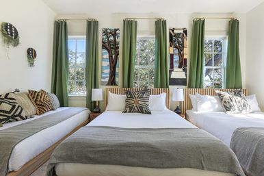 Sunlit guestroom with three low wooden beds, crisp white bedding and gray herringbone throws, patterned accent pillows, green floor-length curtains framing three windows with leafy tree views, wood-slat headboard and woven wall hangings.