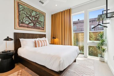 Sunlit modern bedroom with white linens on a wooden platform bed, textured headboard and tree artwork, mustard curtains framing floor-to-ceiling windows with an urban brick view, black bedside lamps and a potted plant.