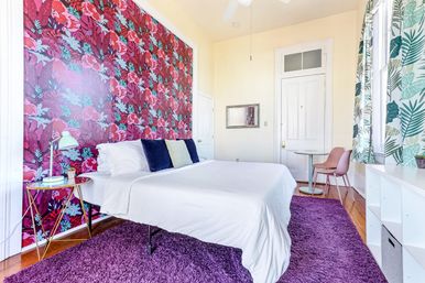 Sunlit bedroom with white bed and linens against a bold pink-red tropical floral accent wall, purple shag rug on hardwood floor, leaf-print curtains at a large window, gold side table with lamp and a small pink dining nook.
