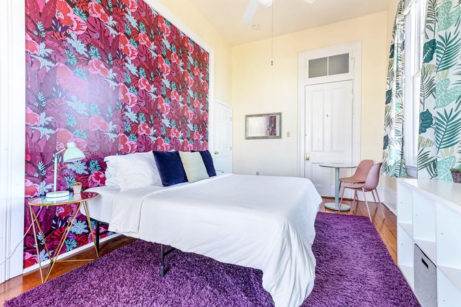 Sunlit bedroom with white bed and linens against a bold pink-red tropical floral accent wall, purple shag rug on hardwood floor, leaf-print curtains at a large window, gold side table with lamp and a small pink dining nook.