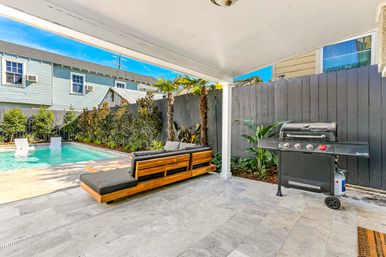 Covered residential backyard patio with teak-style lounge sofa and gray cushions, stone tile floor, wheeled gas grill, and a turquoise swimming pool framed by palm trees and a dark privacy fence on a sunny day