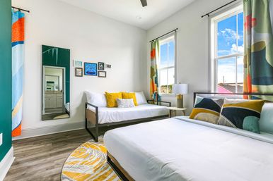 Bright modern guest bedroom with a queen bed and daybed, colorful geometric curtains and accent pillows, large sunlit windows, round yellow rug and hardwood floors.