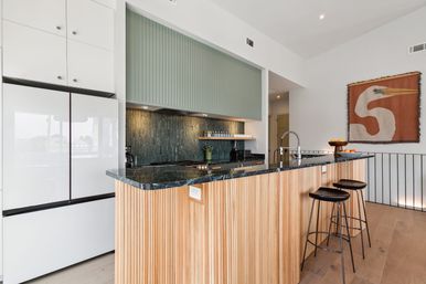 Sunlit modern open-concept kitchen with a ribbed wood island topped by a dark marble countertop, green textured backsplash and cabinets, stainless faucet, two black bar stools and bird wall art.