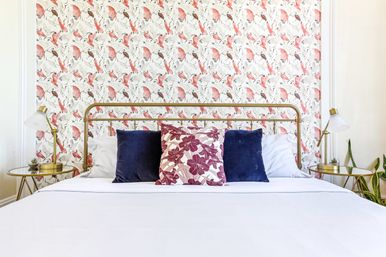 Cheerful modern bedroom with brass bed frame, crisp white bedding, navy and floral throw pillows, pink bird-and-leaf patterned wallpaper, and gold bedside tables with lamps and plants.