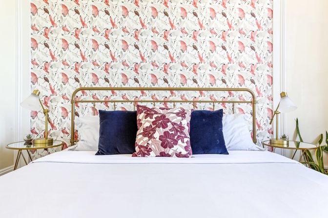 Cheerful modern bedroom with brass bed frame, crisp white bedding, navy and floral throw pillows, pink bird-and-leaf patterned wallpaper, and gold bedside tables with lamps and plants.