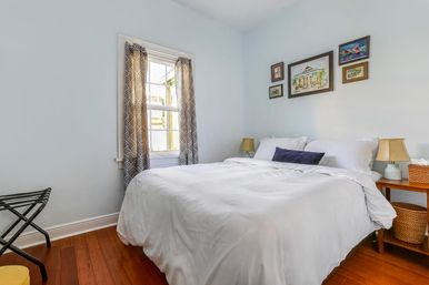 Bright cozy bedroom with white duvet on a queen bed, hardwood floors, patterned gray curtains at a sunny window, framed artwork above the bed, wooden nightstands with lamps and wicker baskets