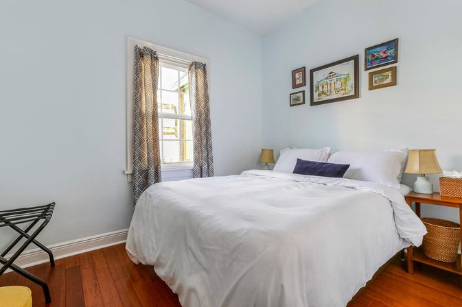 Bright cozy bedroom with white duvet on a queen bed, hardwood floors, patterned gray curtains at a sunny window, framed artwork above the bed, wooden nightstands with lamps and wicker baskets