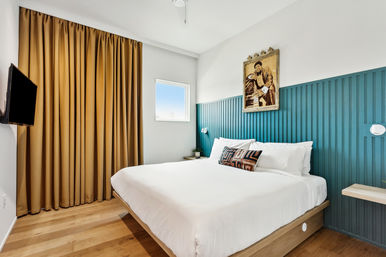 Bright modern boutique hotel bedroom with teal paneled accent wall, king bed dressed in crisp white linens and a patterned lumbar pillow, mustard curtains, hardwood floors, small window and wall-mounted TV.