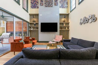 Sunlit double-height modern living room with a large gray sectional facing a wall-mounted TV, built-in wooden shelves, pink accent chair and two rust-orange lounge chairs, striped area rug and brass coffee table, sliding glass doors to a patio with stainless grill and decorative wall art.