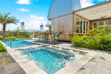 Sunny backyard with bubbling hot tub and adjacent swimming pool, modern wood-clad house patio with outdoor dining, palm tree and steel lift bridge visible in the background.