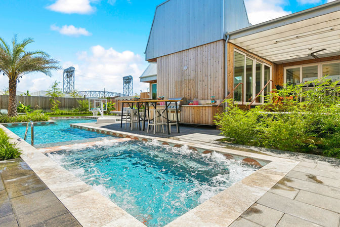 Sunny backyard with bubbling hot tub and adjacent swimming pool, modern wood-clad house patio with outdoor dining, palm tree and steel lift bridge visible in the background.