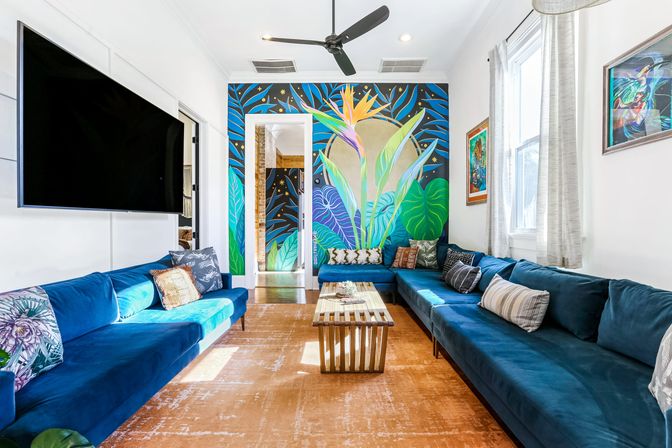 Sunlit modern living room featuring a deep-blue velvet L-shaped sectional, wooden slatted coffee table, wall-mounted TV, ceiling fan, and a vibrant tropical mural accent wall.