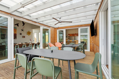 Sunlit enclosed patio dining area with translucent pergola and ceiling fan, wood-clad walls and deck, sliding glass doors to indoor dining and kitchen, black oval tables and green modern chairs.