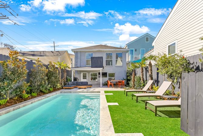 Sunny suburban backyard with a rectangular turquoise swimming pool, artificial turf lawn, three chaise lounges, palm trees, and a modern two-story house with covered patio and outdoor dining area