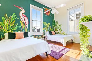 Sunlit tropical mural bedroom with a large heron painted on a green accent wall, two white beds with colorful throw pillows, hardwood floors, purple shag rug, large sash windows, ceiling fan, and trailing indoor plants.